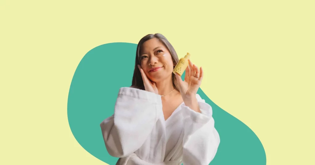 woman in white robe smiling, looking off camera and holding a yellow agency skincare bottle