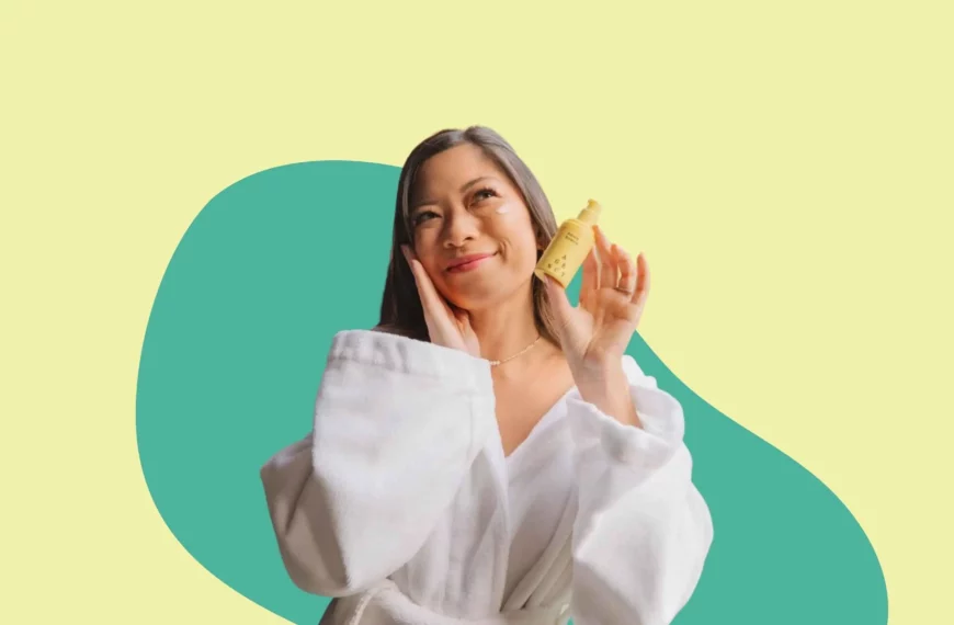 woman in white robe smiling, looking off camera and holding a yellow agency skincare bottle