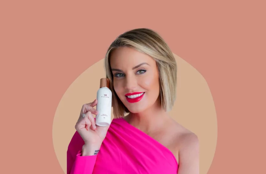 Whitney rose with blonde hair, wearing hot pink dress, holding bottle of wild rose beauty cleanser