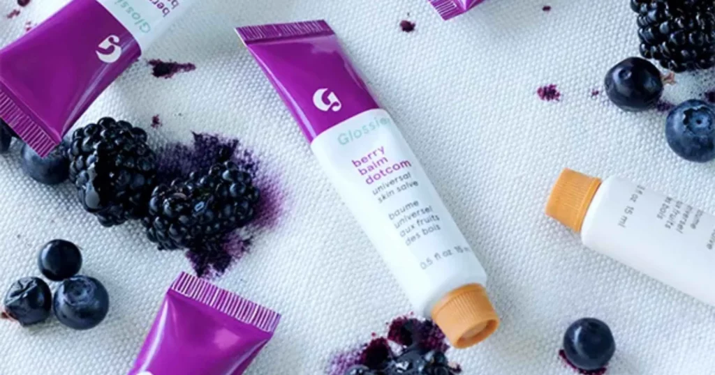 scattered purple and white tubes of Glossier's Balm Dotcom salve with fruit in background
