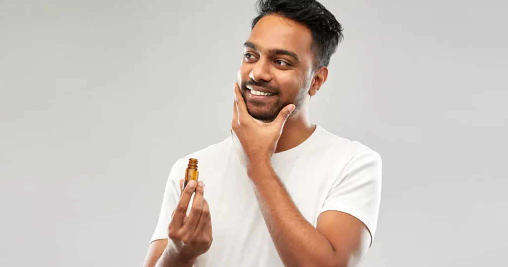 man looking off camera, smiling, touching beard and holding a bottle of beard care product