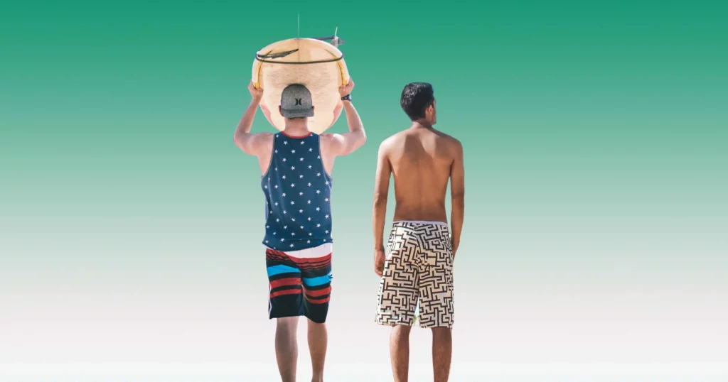 two men turned around, one wearing striped board shorts and blue tank with surfboard on head, the other wearing patterned swim trunks