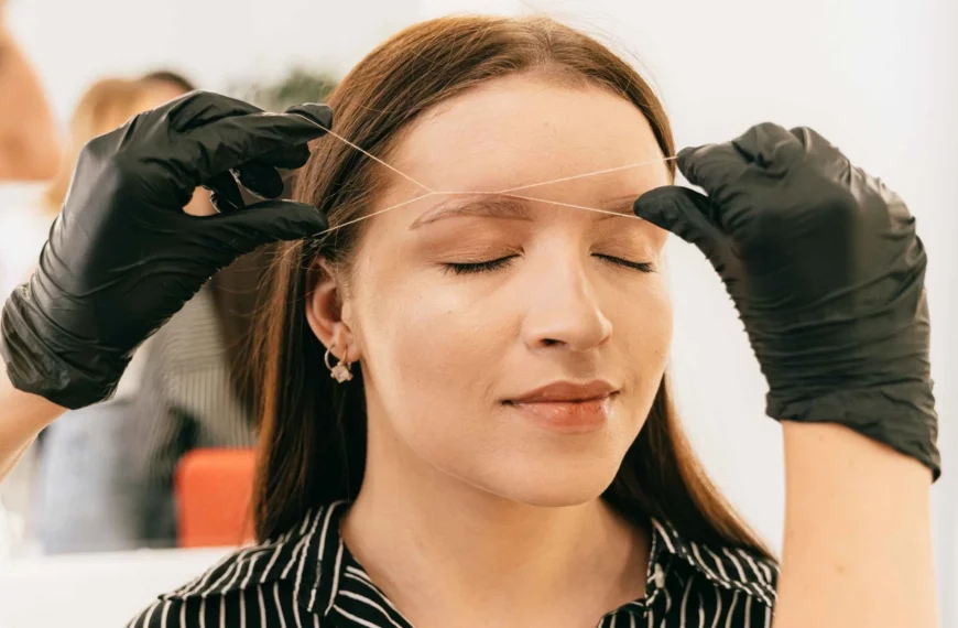 woman with eyes closed having eyebrow threaded by person wearing black gloves