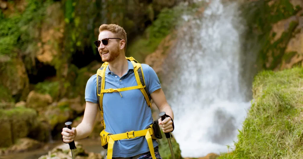 man looking off camera, wearing blue shirt and yellow backpack, holding hiking poles; greenery behind him