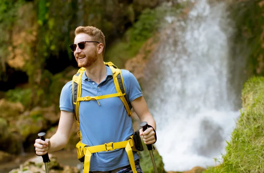 man looking off camera, wearing blue shirt and yellow backpack, holding hiking poles; greenery behind him