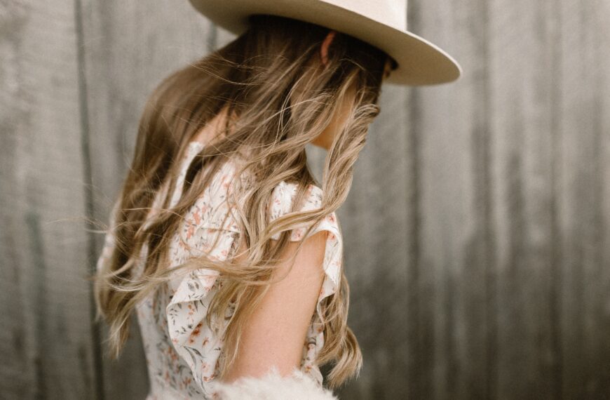 featured image for brands like Doen - this photo shows a close up of the back of a skinny blonde woman wearing a white sun hat and a flowy white dress with floral print. Only her torso and above is visible in the photo.
